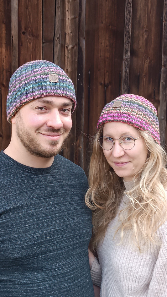 Andreas und Lisa stehen Arm in Arm vor einem Holztor und lächeln in die Kamera. Andreas trägt die Ringelmütze mit vor allem blauen und lila Streifen, Lisa trägt die Variante mit rosa und orange. Beiden passt die Mütze und die Ohren sind bedeckt.