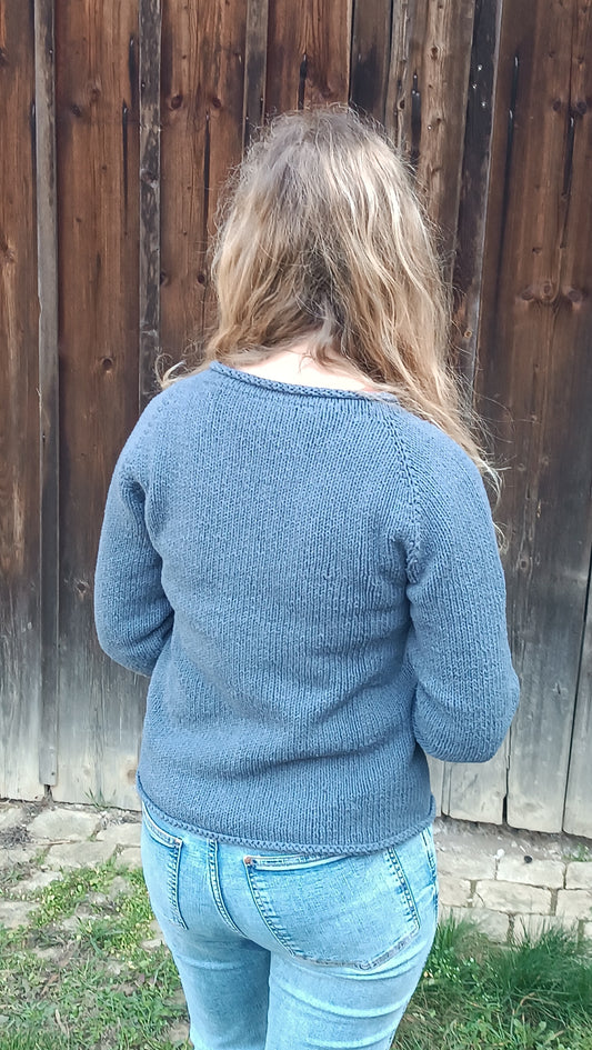 Lisa steht mit dem Rücken zum Betrachter, die offenen Haare über beide Schultern nach vorne gelegt. Sie trägt Jeans und den rauchblauen Strickpullover, die Arme sind abgewinkelt, da sie die Hände versteckt vor dem Körper hält. Durch die Bewegung ergeben sich Bewegungsfalten am Armansatz.