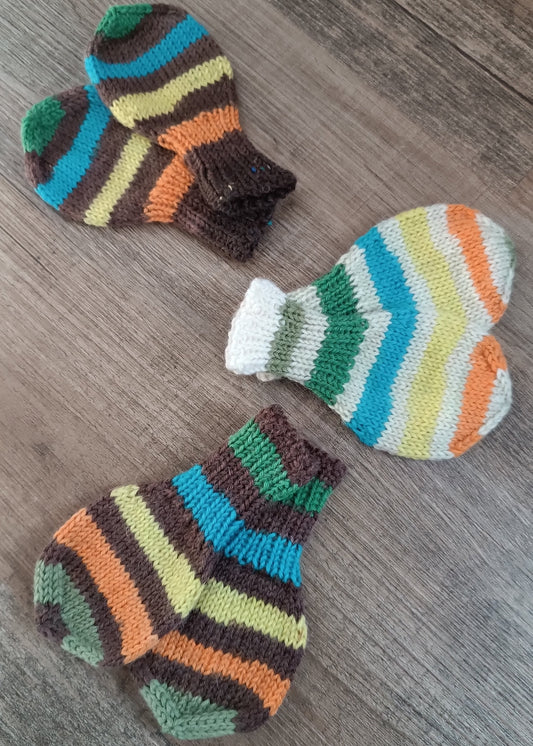 3 Paar Baby-Fausthandschuhe mit bunten Streifen liegen auf einem Holzboden. Jedes Paar zeigt nach außen in eine andere Richtung.