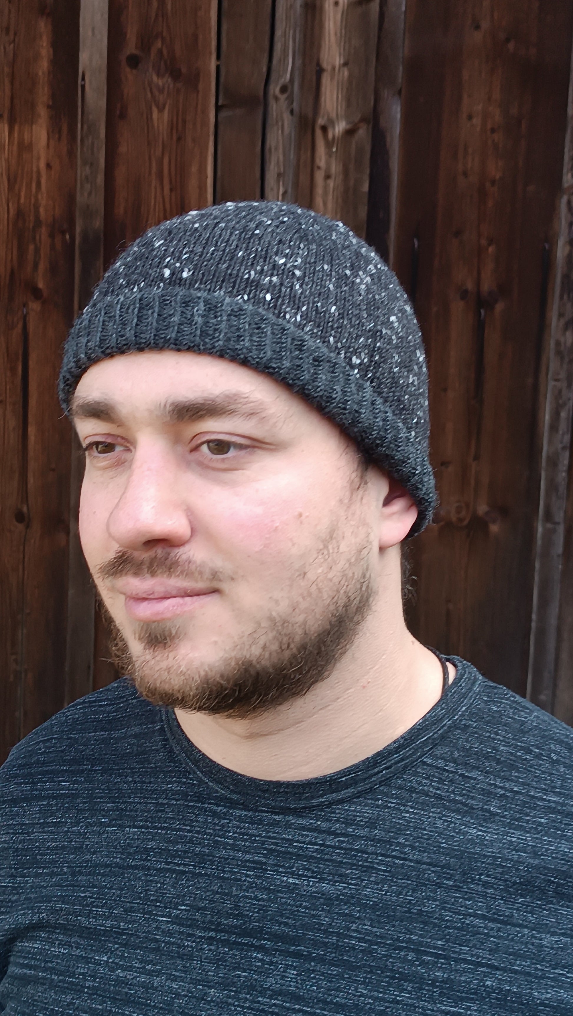 Andreas trägt die graue Beanie mit halb aufgeschlagenem Bündchen und passend dazu ein graues T-Shirt. Er steht vor einem braunen Holztor und blickt gedankenverloren links aus dem Bild.