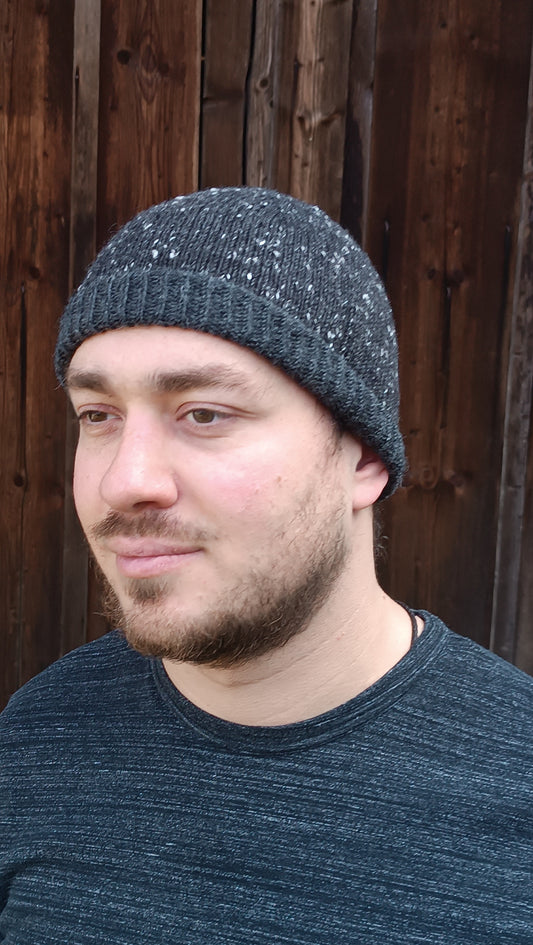 Andreas trägt die graue Beanie mit halb aufgeschlagenem Bündchen und passend dazu ein graues T-Shirt. Er steht vor einem braunen Holztor und blickt gedankenverloren links aus dem Bild.