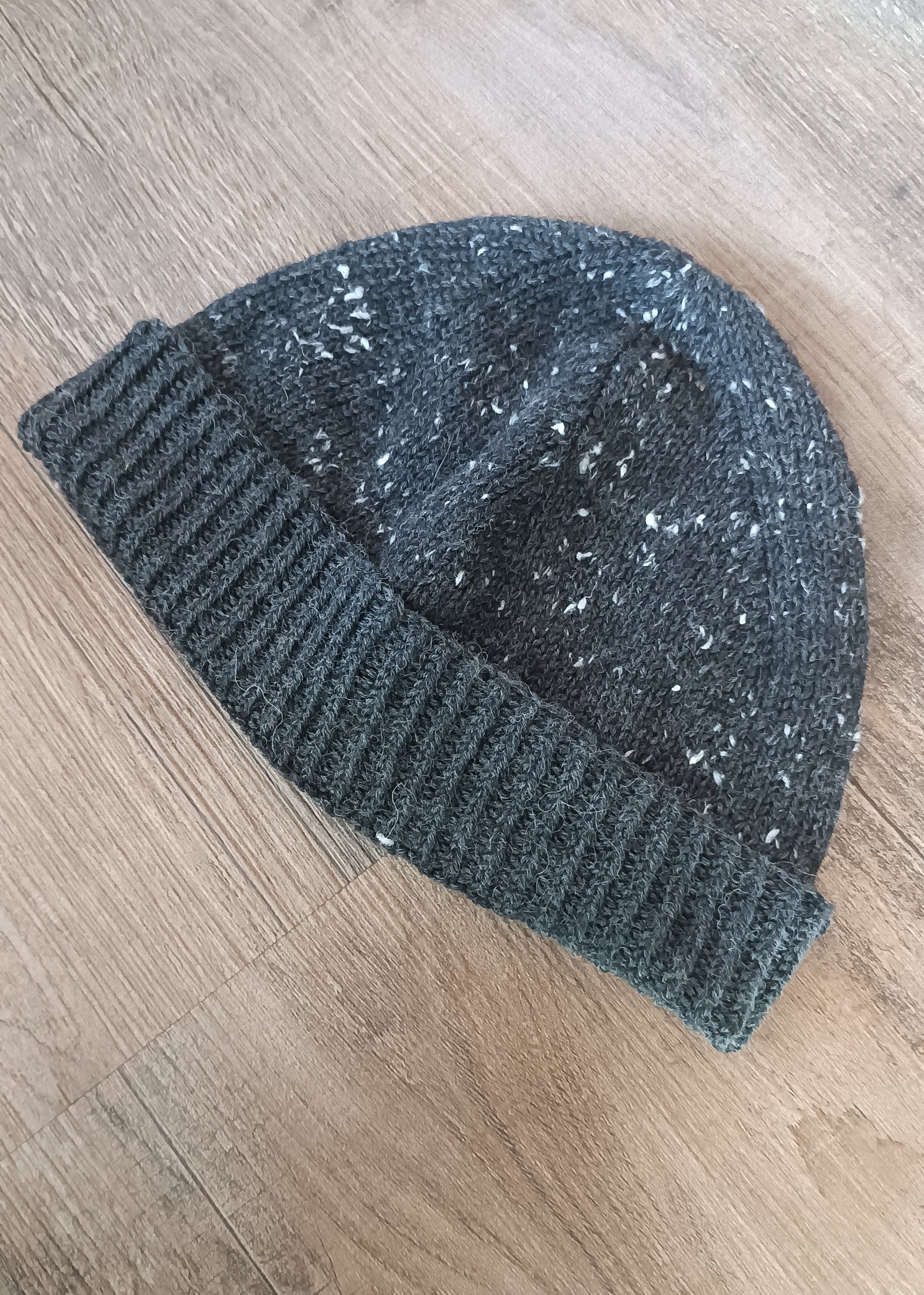Eine anthrazitfarbene Beanie mit Tweed-Noppen liegt schräg im Bild, das 1/1-Rippenbündchen ist nach oben geschlagen.