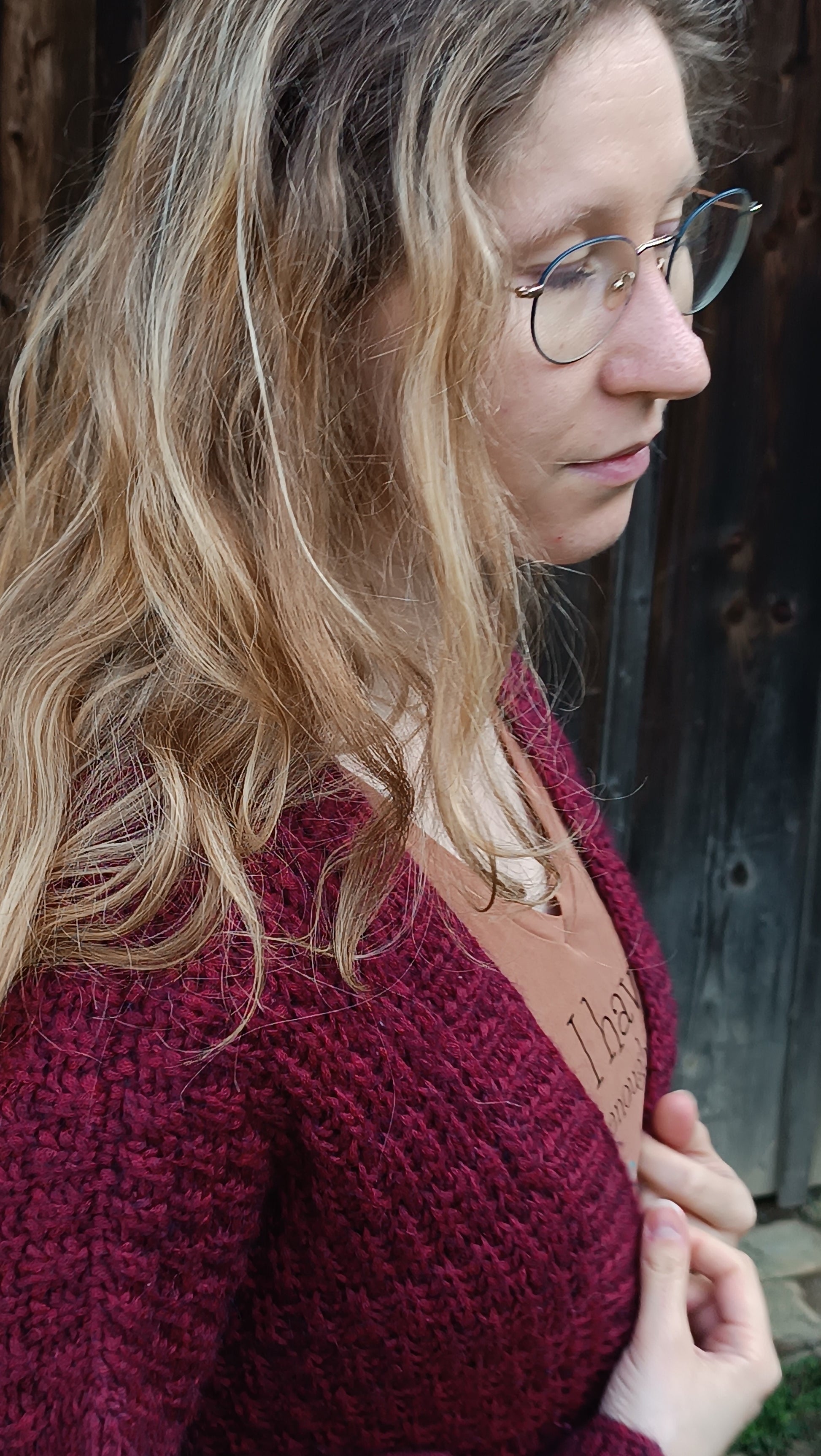 Lisa steht mit geschlossenen Augen und offenen Haaren nach rechts gewandt im Bild. Sie trägt ein braunes T-Shirt und eine weinrote Strickjacke. Das unterbrochene oder verschobene Rippenmuster des Cardigans sowie die Schulternaht ist gut zu sehen.