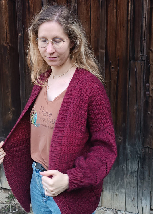 Lisa steht mit geschlossenen Augen und offenen Haaren vor einem Holztor. Ihr Körper ist leicht zur linken Bildseite gewendet. Sie trägt ein braunes T-Shirt, Jeans und den weinroten Strick-Cardigan. Sie hält ihn mit beiden Händen vorne fest, als würde sie ihn gerade um sich schlingen wollen.