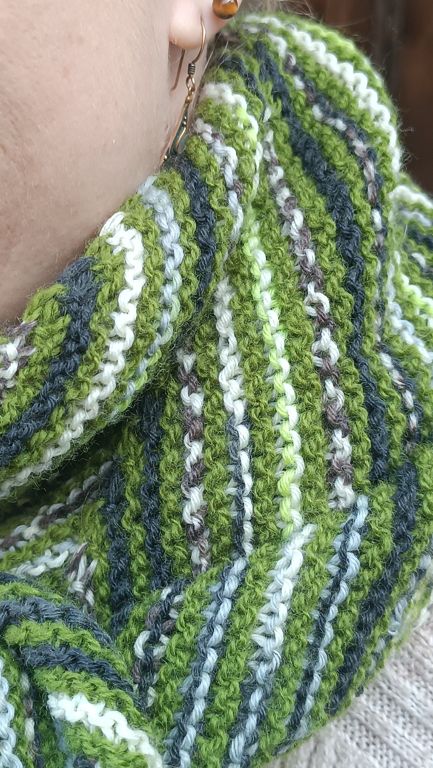 Eine Nahaufnahme des gestreiften Dreieckstuch. Die Rippen des kraus rechts gestrickten Tuchs sind zu erkennen, ebenso die Abfolge des einfarbig grünen Garns sowie des gemusterten zweiten Garns.