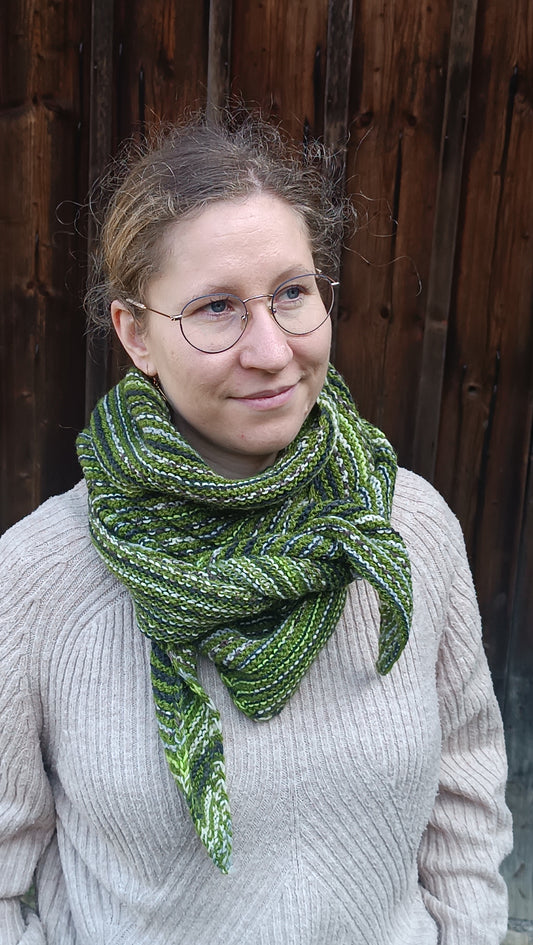 Lisa steht vor einem Holztor und schaut schräg nach rechts aus dem Bild. Sie trägt einen beigen Strickpullover und das gestreifte Dreieckstuch. Die Enden sind dieses Mal vorne über der mittleren Spitze miteinander verknotet.