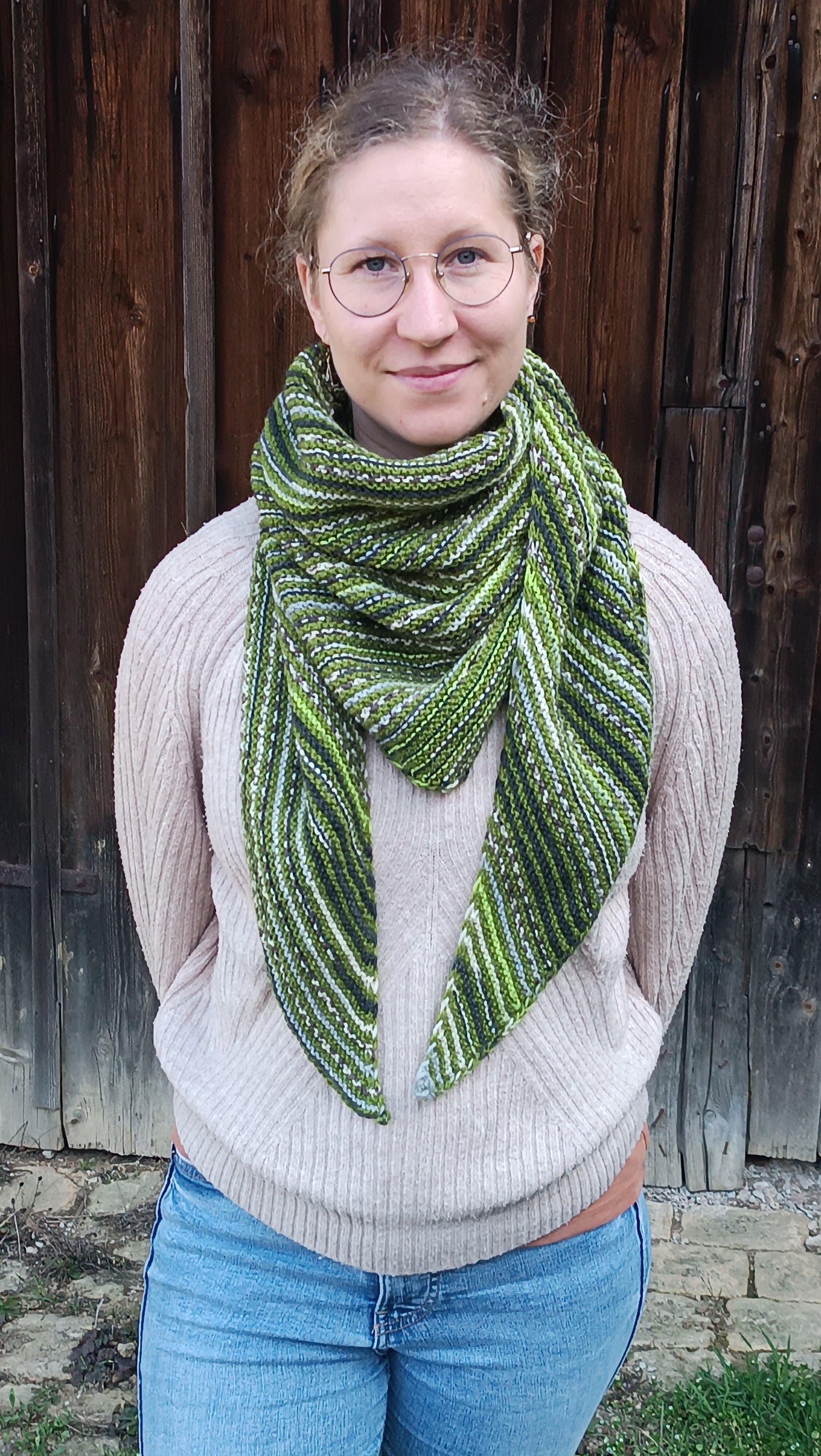 Lisa steht mit den Armen auf dem Rücken vor einem Holztor und schaut lächelnd zur Kamera. Sie trägt Jeans, einen beigen Strickpullover und das gestreifte Dreieckstuch in grün, weiß, grau und schwarz. Die Enden hängen locker nach unten und reichen etwa bis zum Bauchnabel.