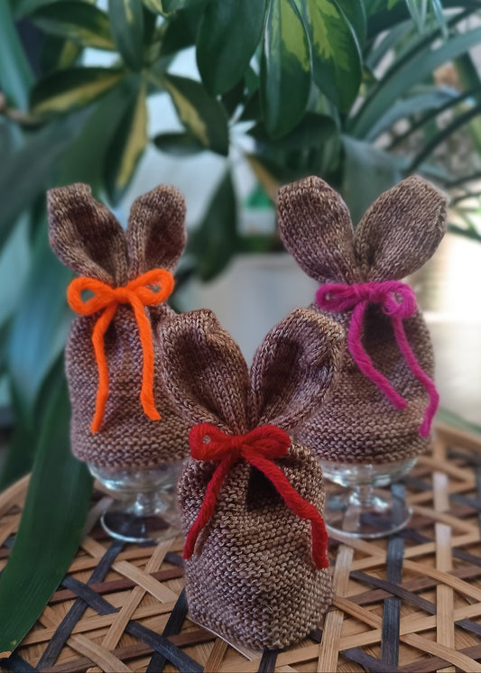 Drei braune Eierwärmer mit Hasenohren und bunten Schleifen stehen auf einem geflochtenen Untergrund vor einigen Pflanzen.