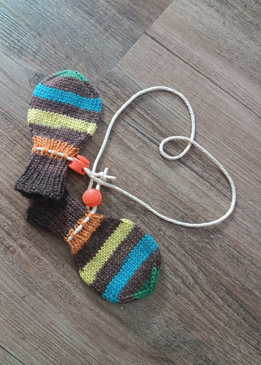 Ein Paar gestreifte Fäustlinge liegt auf dem Boden. Die beiden sind mit einer Naturfarbenen Kordel verbunden, die neben den Fäustlingen ein Herz bildet.