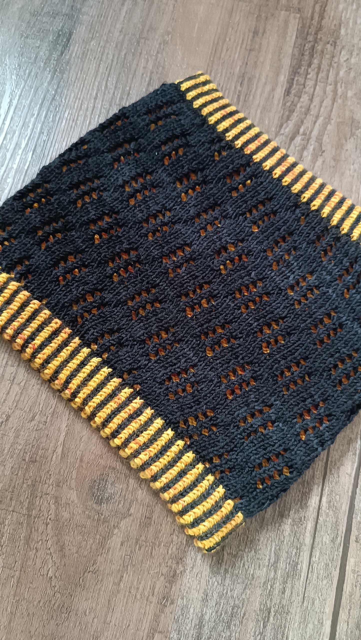 Das Bild zeigt einen handgestrickten Cowl, die äußere Lage ist aus schwarzem Garn mit einem Lochmuster. Darunter sieht man eine Lage in gelb. Oben und unten ist ein zweifarbiges Bündchen aus gelbem und schwarzem Garn.