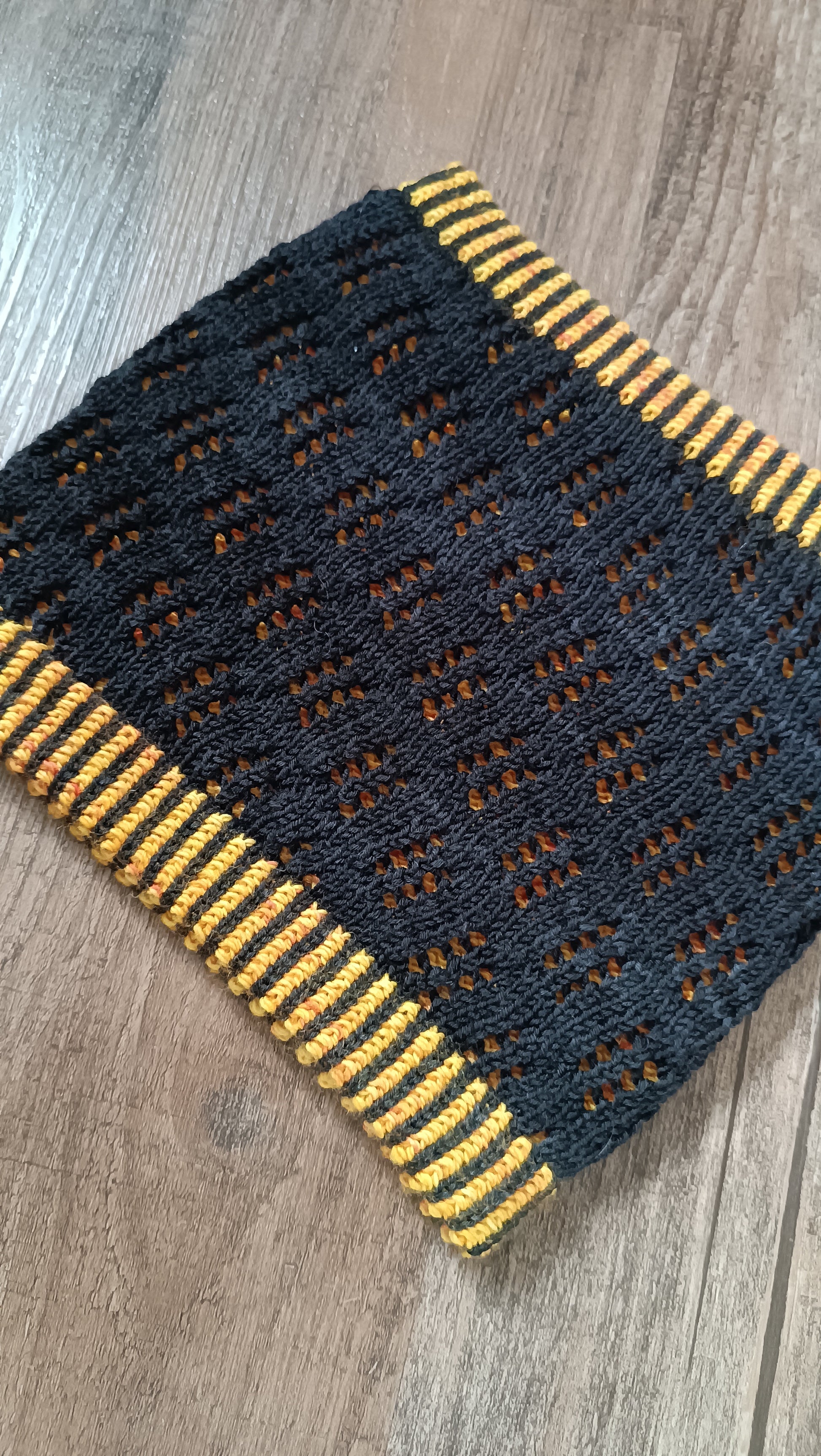 Das Bild zeigt einen handgestrickten Cowl, die äußere Lage ist aus schwarzem Garn mit einem Lochmuster. Darunter sieht man eine Lage in gelb. Oben und unten ist ein zweifarbiges Bündchen aus gelbem und schwarzem Garn.
