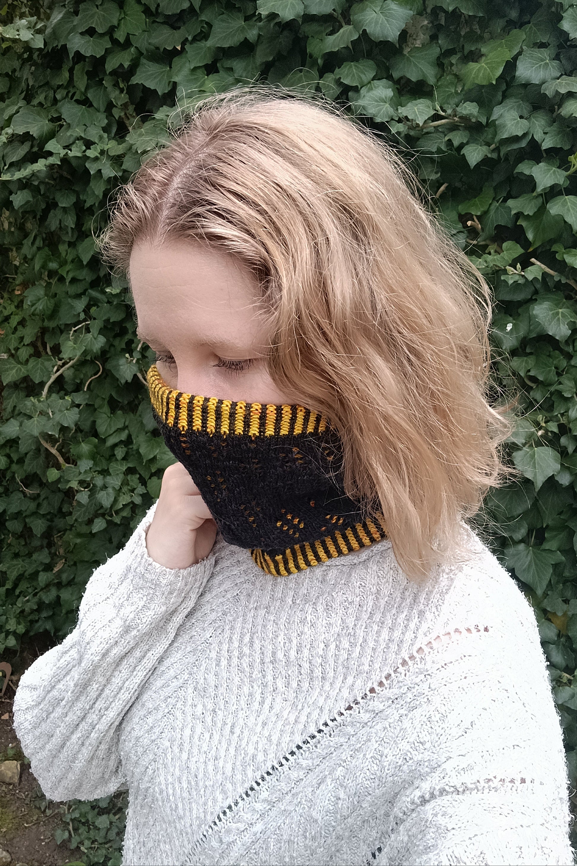 Lisa steht seitlich vor einer Efeu-Wand. Sie trägt einen halsnahen, handgestrickten Cowl in schwarz und gelb, den sie bis über die Nase nach oben gezogen hat. Er reicht unten bis zum Schlüsselbein. Dazu trägt sie die blonden Haare offen und einen hellgrauen Strickpulli.