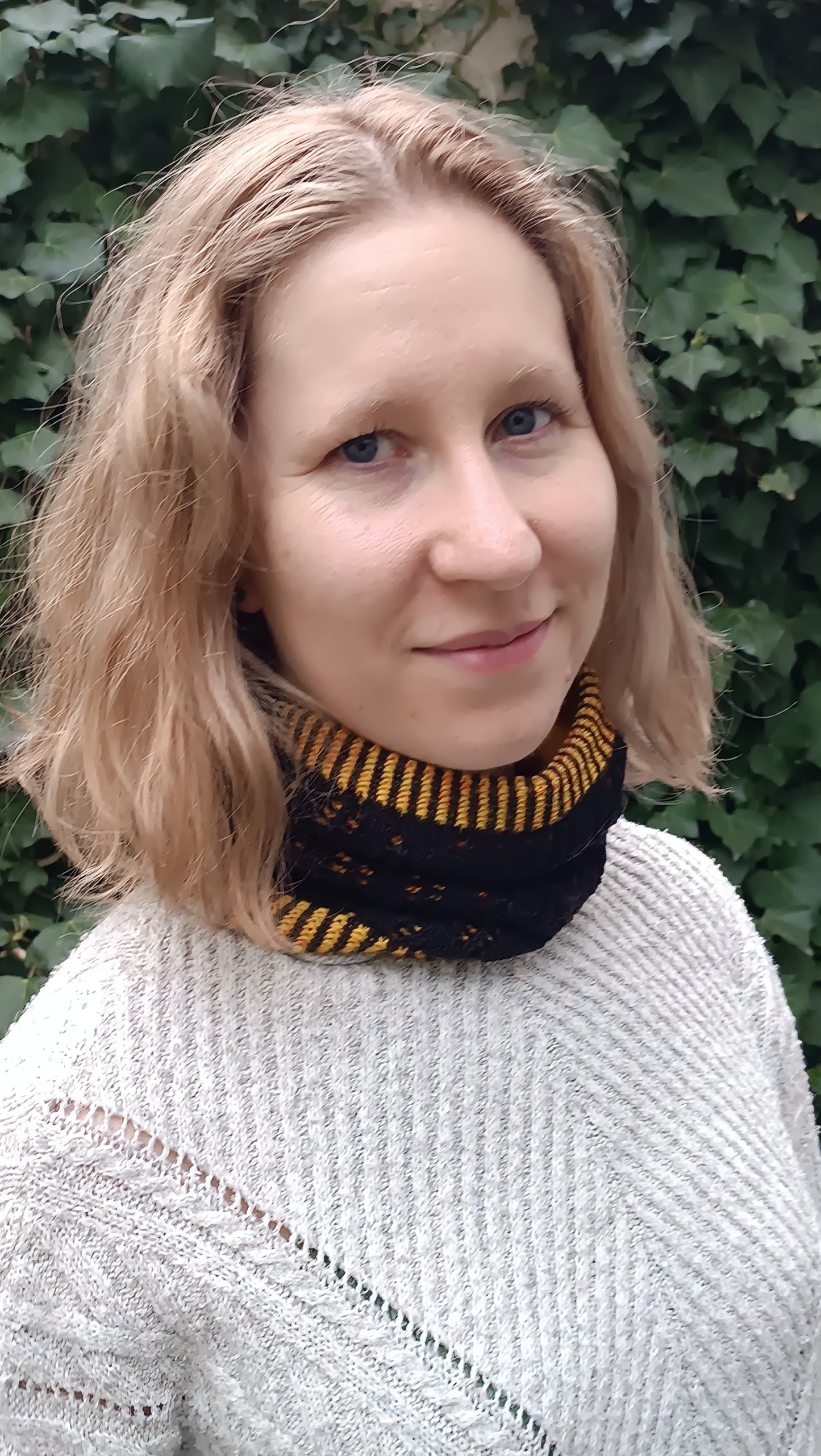 Lisa steht leicht seitlich vor einer Efeu-Wand. Sie trägt einen schwarz-gelben, handgestrickten Cowl. Dazu einen hellgrauen Strickpulli und ihre blonden, schulterlangen Haare offen. Sie lächelt leicht.