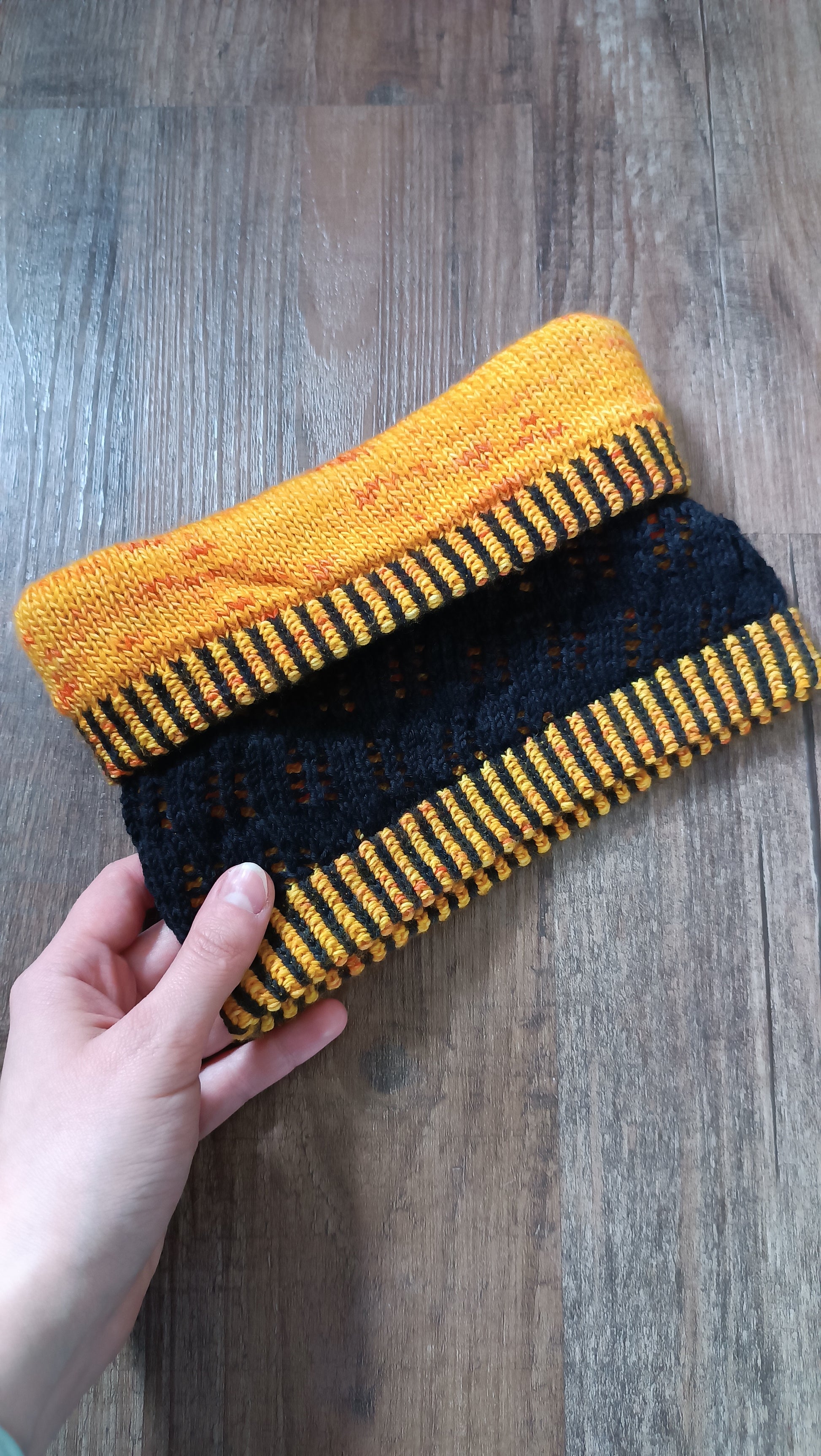 Eine hellhäutige Hand hält von links einen handgestrickten Cowl ist Bild. Die obere Kante ist nach unten geklappt, circa zu einem Drittel. So sieht man oben die gelbe Innenseite, dann das zweifarbige Bündchen, darunter die schwarze Außenseite mit Lochmuster und unten wieder ein zweifarbiges Bündchen.