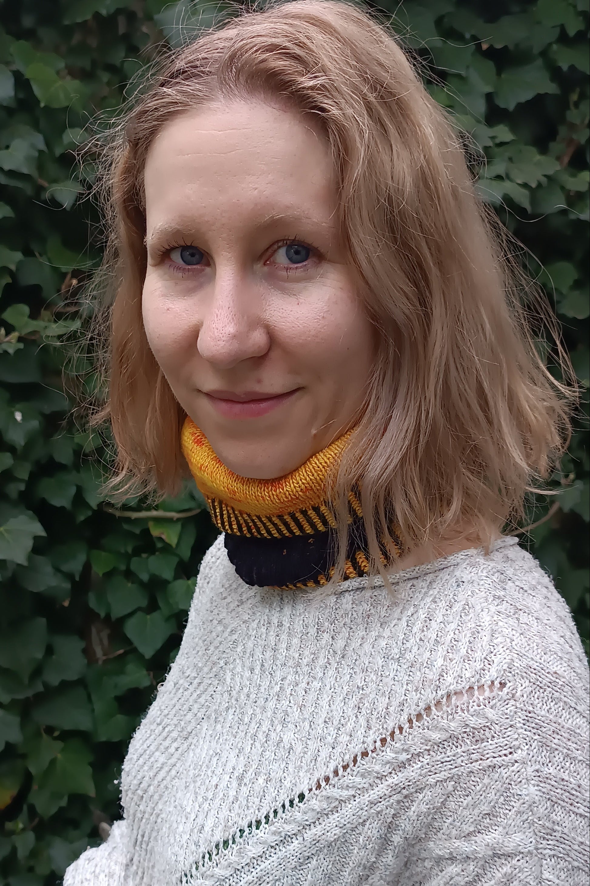 Lisa steht seitlich vor einer Efeu-Wand. Sie trägt einen handgestrickten, schwarz-gelben Cowl, dessen oberes Ende nach unten geklappt ist. Dadurch sieht man sowohl die äußere schwarze Lage als auch die innere gelb-orangene Lage des Cowls. Dazu hat sie die blonden, schulterlangen Haare offen und trägt einen hellgrauen Strickpulli.