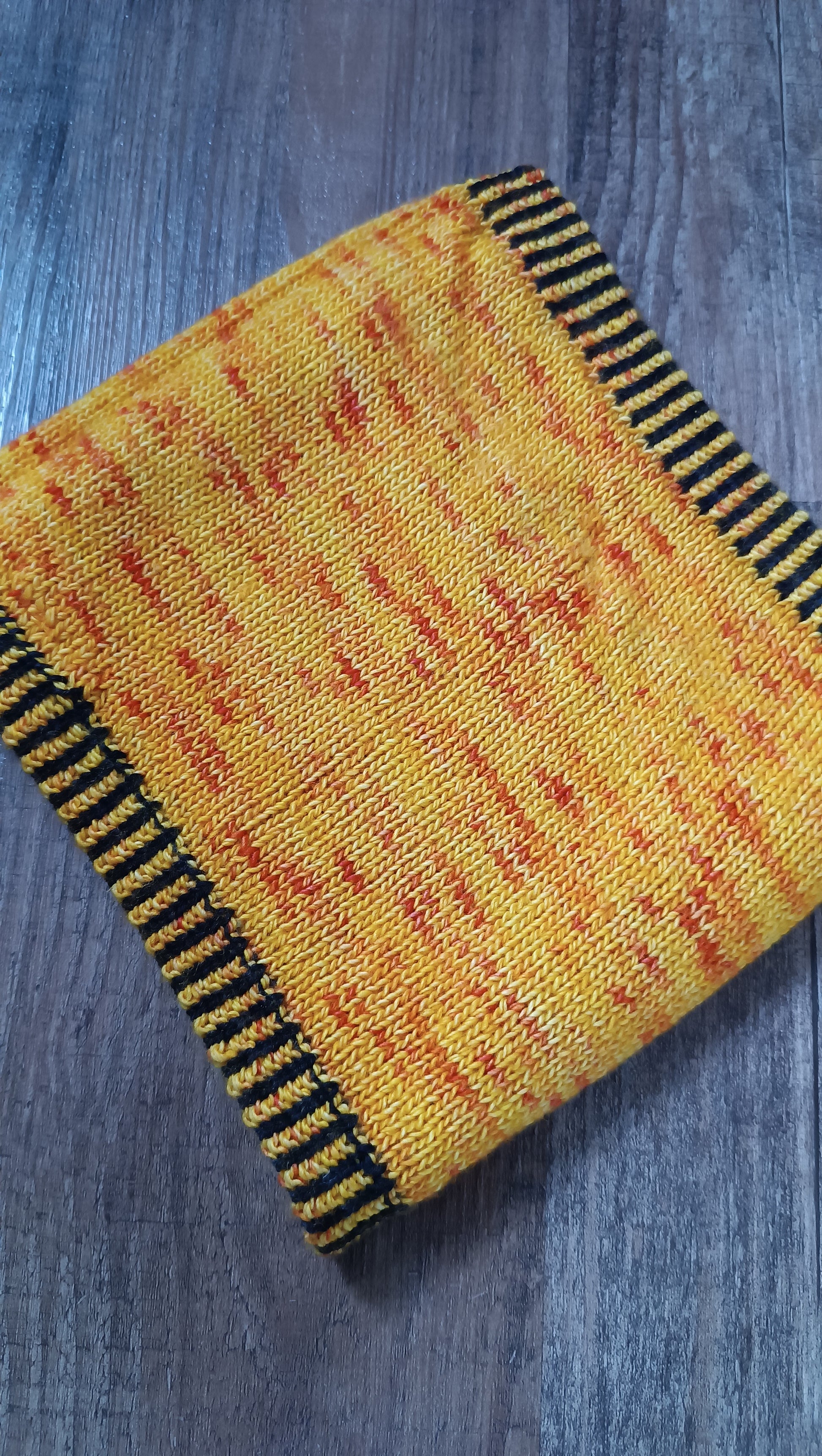 Ein handgestrickter Cowl aus gelbem Garn mit orange-roten Sprengseln und einem zweifarbigen Bündchen in schwarz und gelb. Er liegt schräg im Bild auf einem Holzboden.