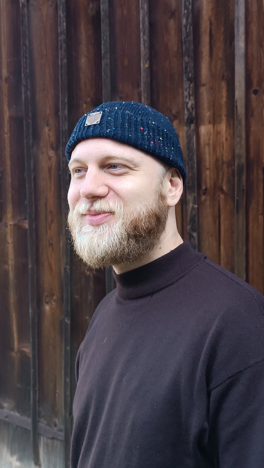 Jonas trägt die nachtblaue Fischermütze zu einem dunkelbraunen Rollkragenshirt und Vollbart. Er steht vor einem Holztor und schaut lächelnd nach linsk aus dem Bild. Die Mütze bedeckt gerade so den oberen Teil seiner Ohren.