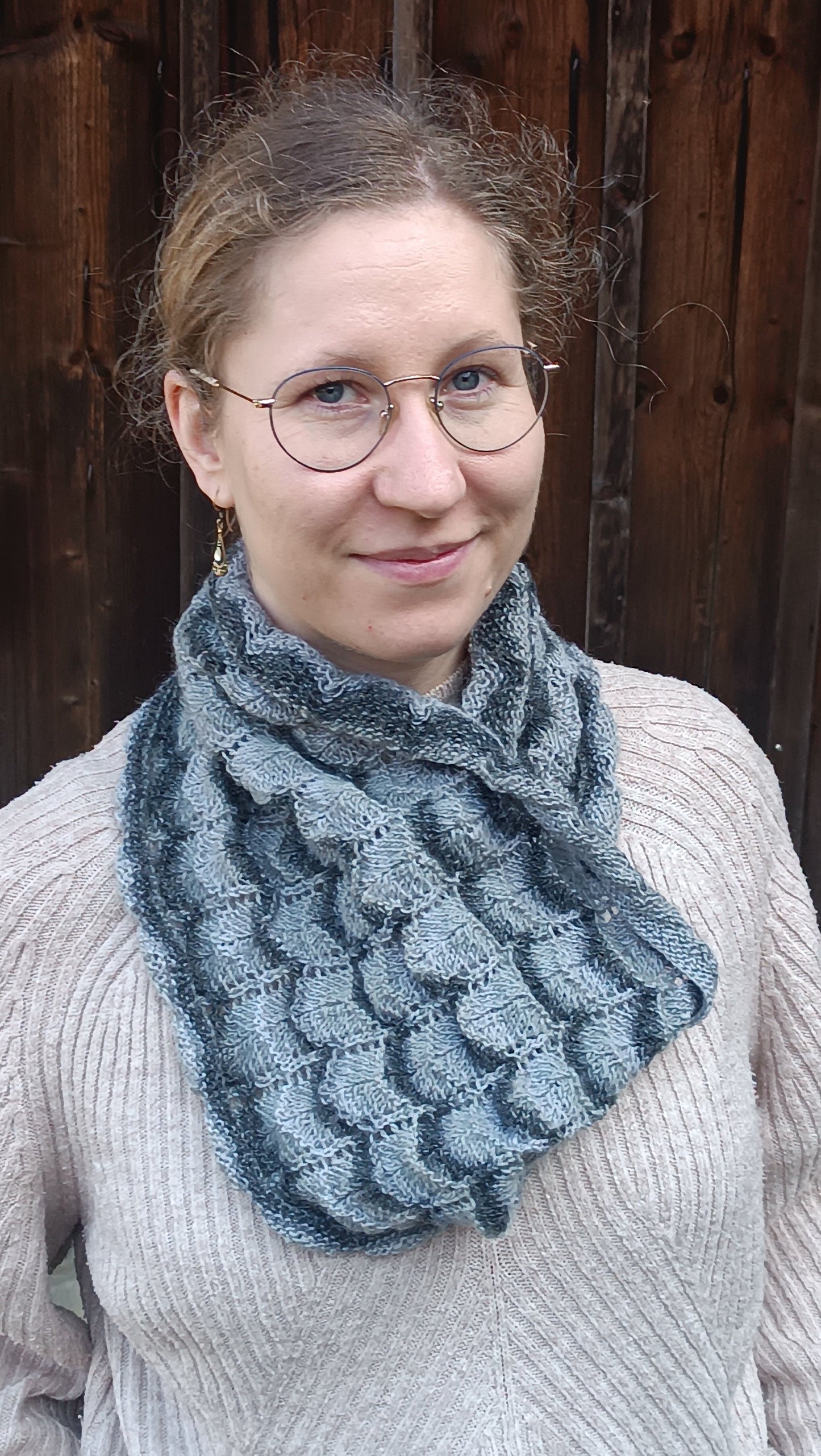 Lisa steht vor einem alten Holztor und lächelt in die Kamera. Sie trägt einen weiten Cowl, der ein Wellenmuster aus mehreren Grautönen und Lochmuster-Streifen zeigt. Er ist auf der Brust schräg gelegt, wie man ihn unter einer Jacke mit tieferem Ausschnitt tragen würde.
