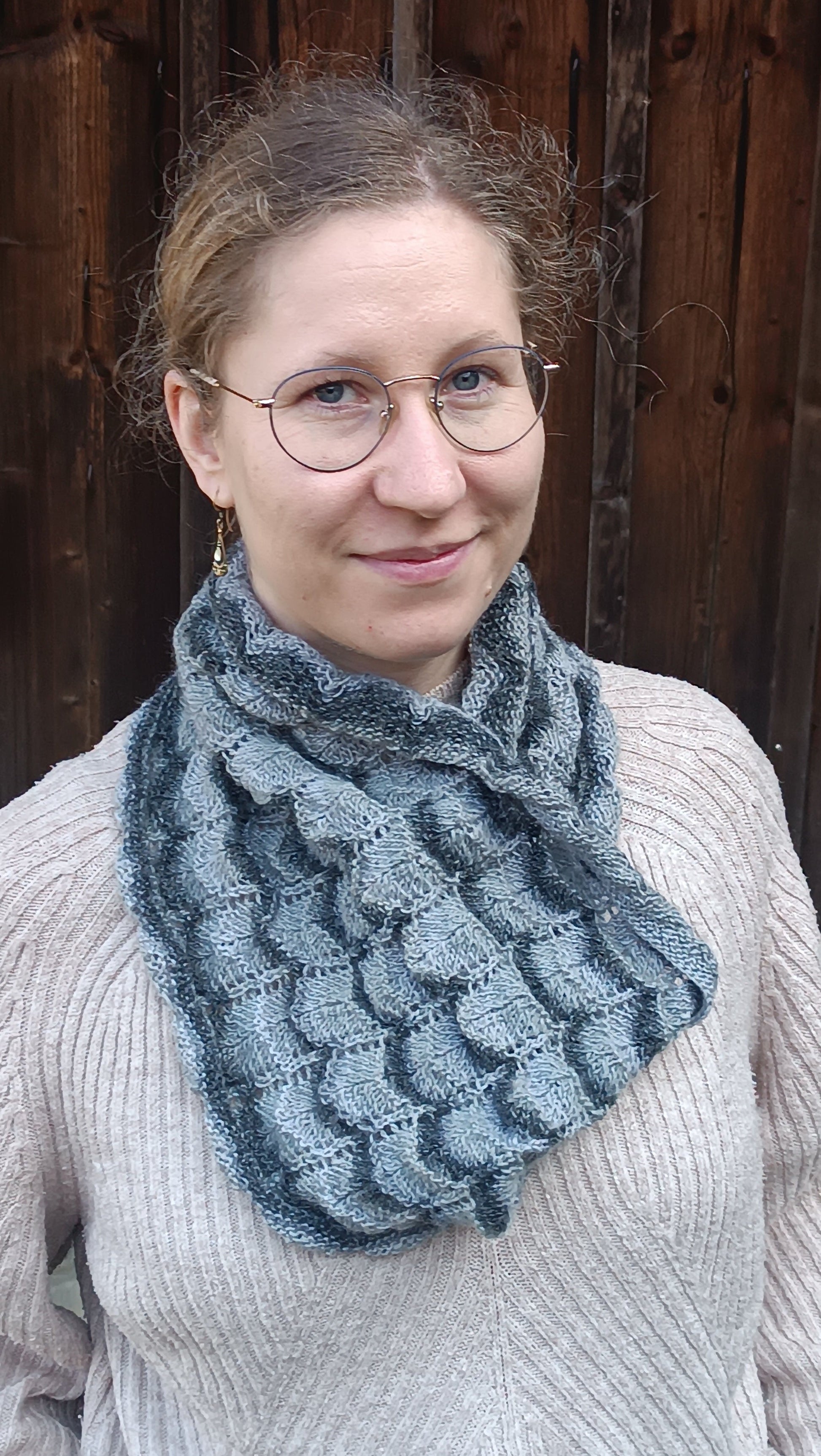 Lisa steht vor einem alten Holztor und lächelt in die Kamera. Sie trägt einen weiten Cowl, der ein Wellenmuster aus mehreren Grautönen und Lochmuster-Streifen zeigt. Er ist auf der Brust schräg gelegt, wie man ihn unter einer Jacke mit tieferem Ausschnitt tragen würde.
