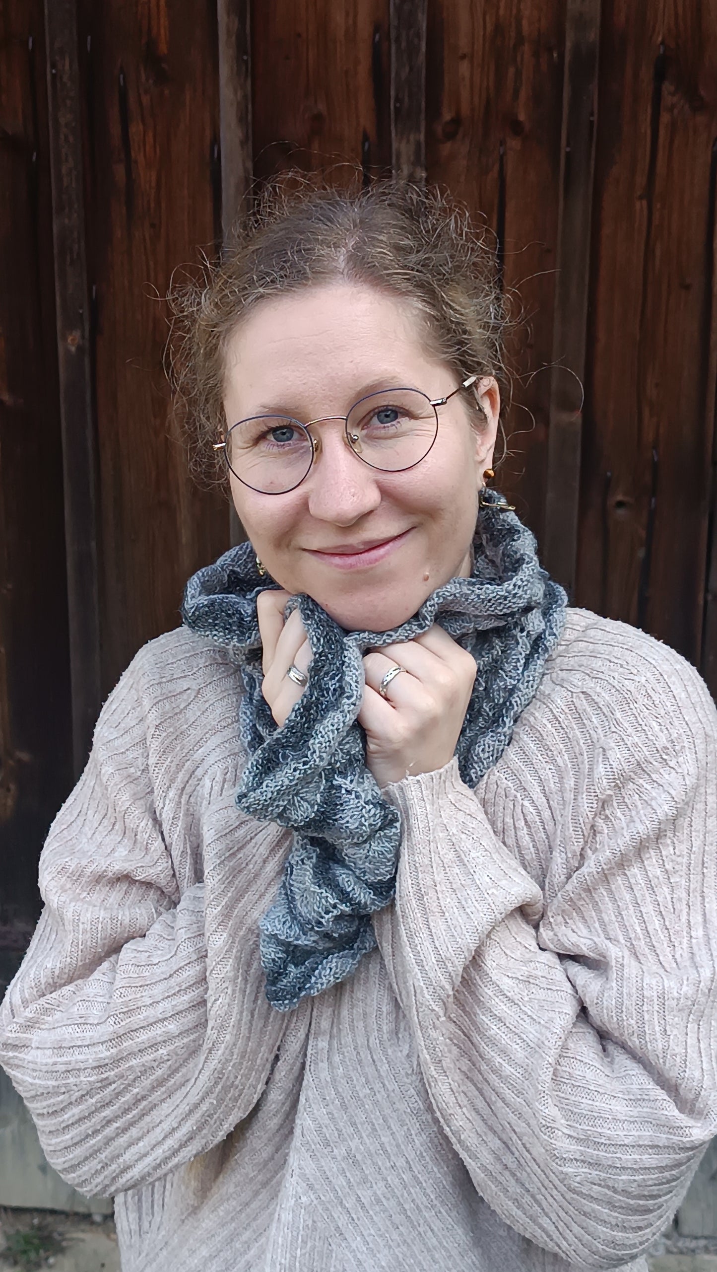Lisa steht vor einem alten Holztor und lächelt in die Kamera. Sie trägt einen weiten, grauen Loopschal, den sie mit beiden Händen an den Hals drückt, um sich darin einzukuscheln. Dazu trägt sei einen beigen Strickpulli.