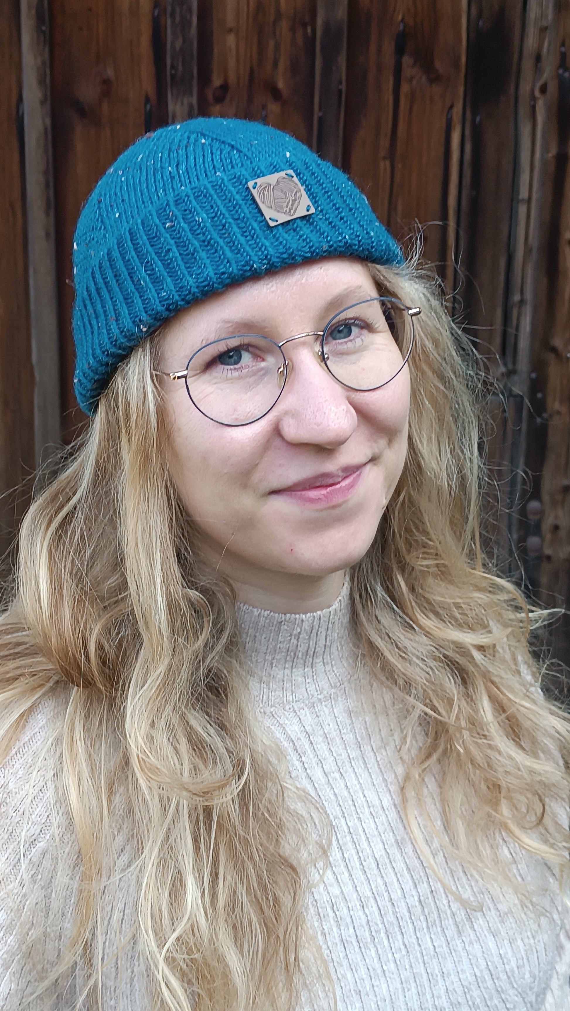 Lisa trägt die Fischermütze in petrol über offenen Haaren. Sie trägt außerdem einen beigen Strickpullover, steht vor einem Holztor und grinst mit schief gelegtem Kopf in die Kamera. 