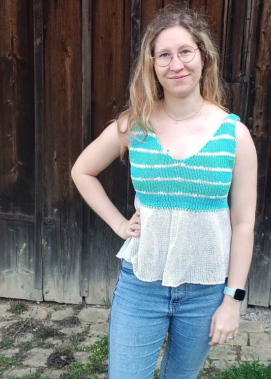 Lisa steht vor einem Holztor, die rechte Hand in der Hüfte, die linke nach unten hängend und grinst in die Kamera. Sie trägt Jeans und ein Top mit V-Ausschnitt. Es hat Querstreifen an den Trägen und im Brustbereich, darunter fällt ein weißes Schößchen etwas ausgestellt nach unten.