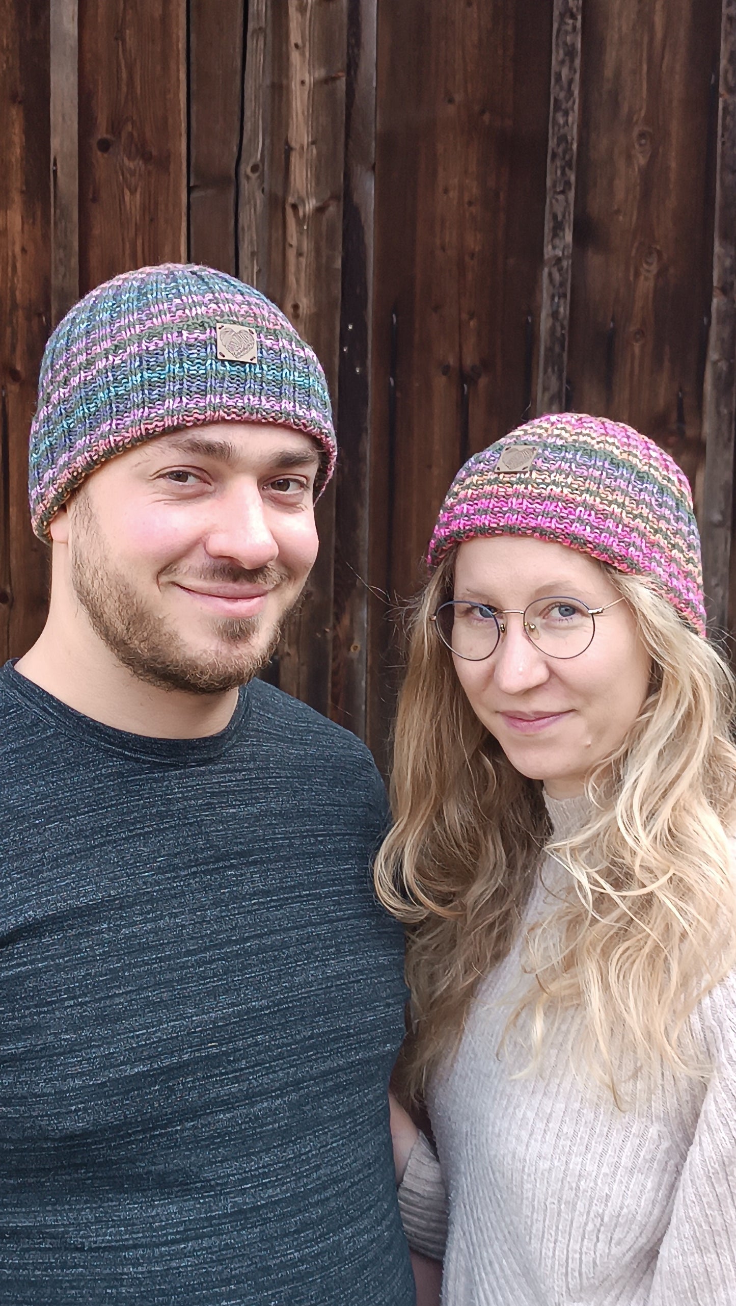 Andreas und Lisa stehen Arm in Arm vor einem Holztor und lächeln in die Kamera. Andreas trägt die Ringelmütze mit vor allem blauen und lila Streifen, Lisa trägt die Variante mit rosa und orange. Beiden passt die Mütze und die Ohren sind bedeckt.