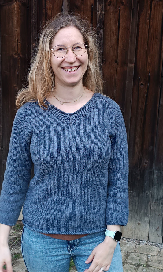 Lisa steht strahlend mit offenen Haaren vor einem Holztor. Sie trägt Jeans und den rauchblauen V-Neck-Sweater, unter dem unten ein braunes T-Shirt hervorblitzt. Der tiefste Punkt des V-Ausschnitts ist ca. eine Handbreit vom Halsansatz entfernt. Der Pullover endet am oberen Ende der Hüfte, die Ärmel haben etwa dieselbe Länge.